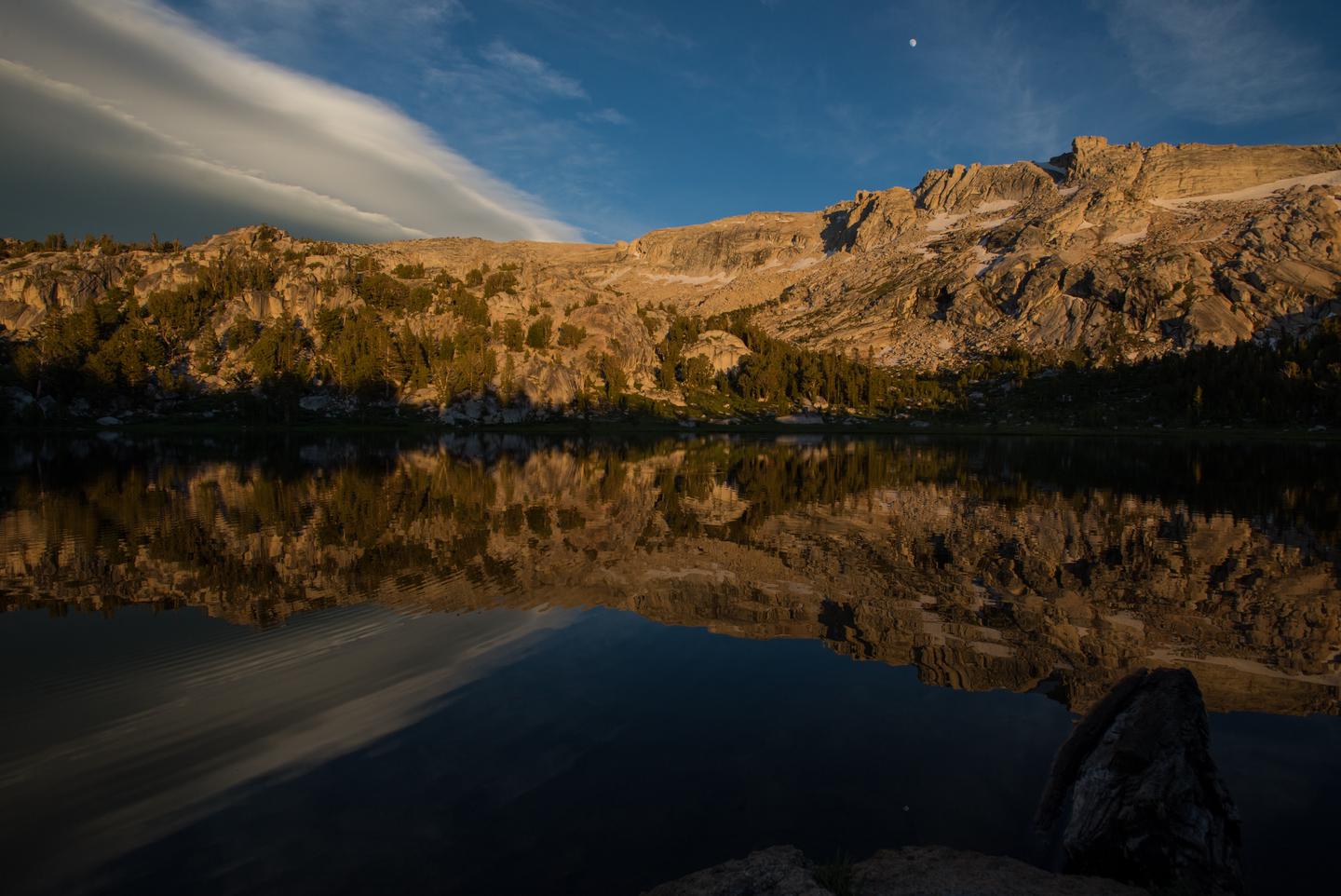 View of lake at sunset, with a mountain behind in alpenglow and the nearly full moon in the sky above.Alpenglow meets the moon, illuminating peaks surrounding a High Sierra lake. 