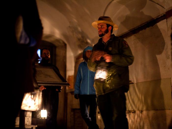 Ranger and visitors stand in the dark with illuminated lanterns casting light and shadows.Ranger and  park visitors stand inside of Fort Point in the dark wearing jackets. They hold all hold lanterns which illuminate the dark halls casting an array of shadows around the brick walls.