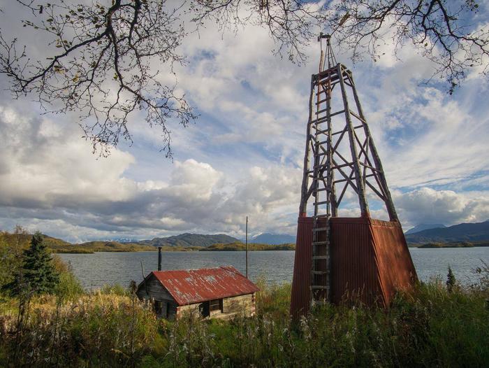 View of a wood cabin and windmill in a grassy field with a lake and mountains in the background.Fure's Cabin and windmill, located in the Bay of Islands, Naknek Lake.