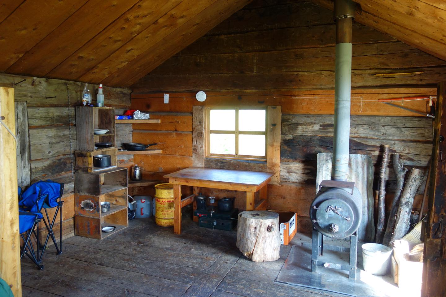 Inside a rustic wooden cabin with a small table, wood burning stove, and small shelves with cooking items.Fure's Cabin has a wood burning stove and space for cooking.