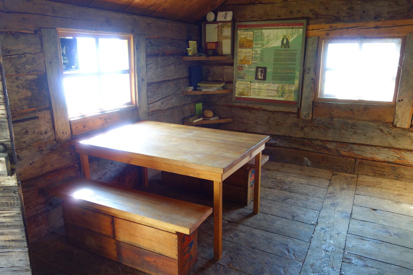 A wooden table and bench sit next to a wall with windows and an informational sign.Fure's Cabin has a table and benches and information about Roy Fure.