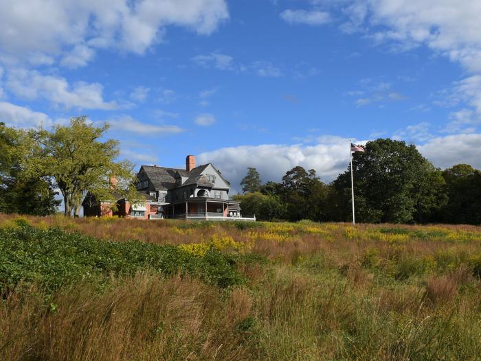 A gray house and flagpole atop a grassy hill with blue sky and clouds in the background.The Roosevelt Home atop Sagamore Hill.