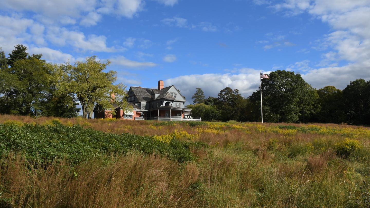 A gray house and flagpole atop a grassy hill with blue sky and clouds in the background.Theodore Roosevelt's home atop Sagamore Hill.