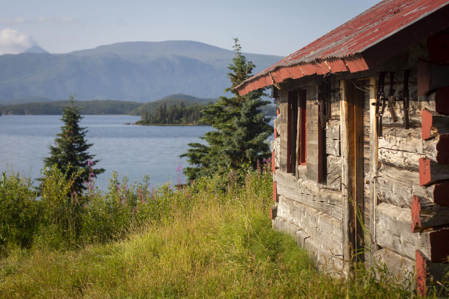 A wood cabin bathed in sunlight sits on a grassy field next to a lake surrounded by mountains.Fure's Cabin sits in a clearing in the Bay of Islands on Naknek Lake.