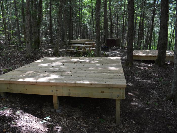 Three wooden tent platforms and two picnic tables, a metal fire pit and food storage locker surrounded by trees at Lunksoos Campsite Seven.Lunksoos Campsite Seven accommodates groups in a shady setting surrounded by trees.
