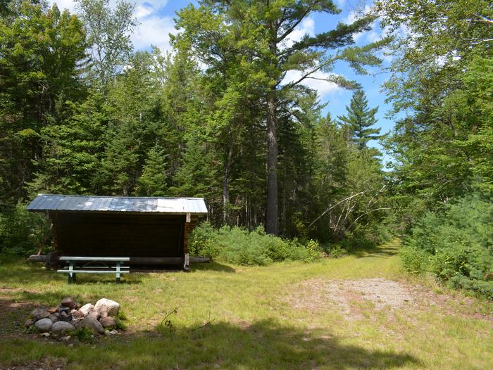 Three-sided wooden shelter, wooden picnic table and stone fire ring at Wassataquoik Lean-to.The Wassataquoik Lean-to offers a sheltered camping experience approximately one mile from the Wassataquoik Gate.