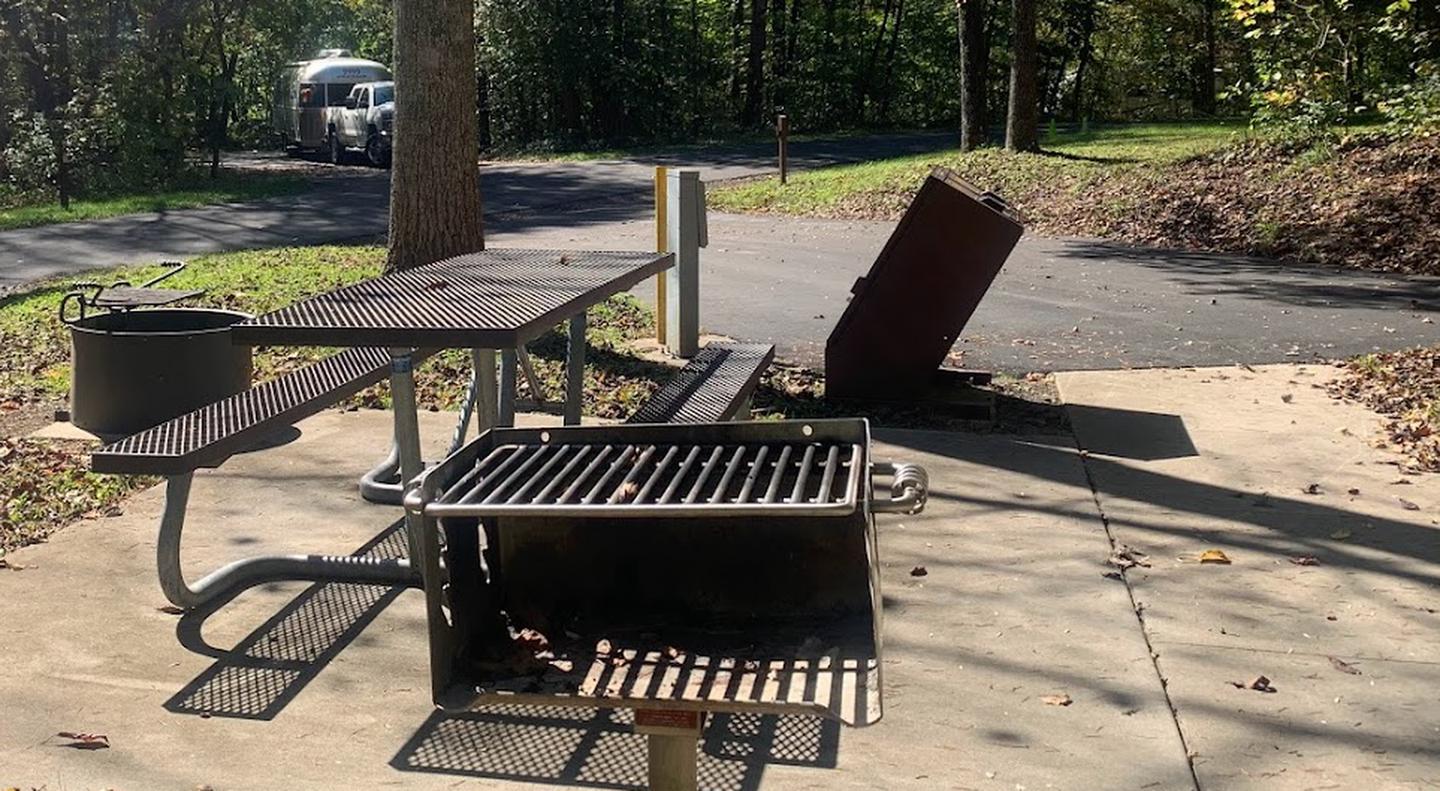 A concrete area with a grill, picnic table, circle fire ring, and a brown bear proof trash can with an under handle push lift.B-6 site with fire ring, grill, and picnic table.
