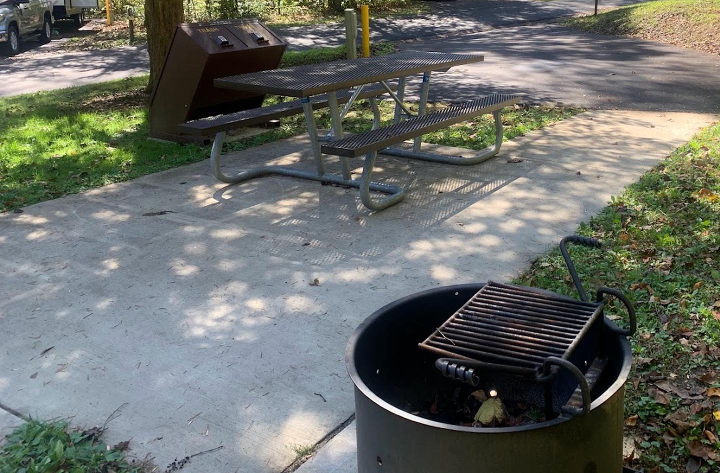 A concrete area with a grill, picnic table, circle fire ring, and a brown bear proof trash can with an under handle push lift.B-8 has a fire ring, grill, and picnic table.