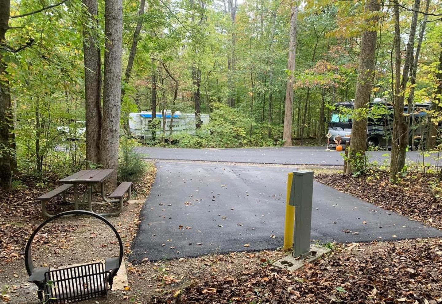 A blacktop surface with a yellow post and gray box near a circle fire ring and picnic table.C-14 has a fire ring and picnic shelter.