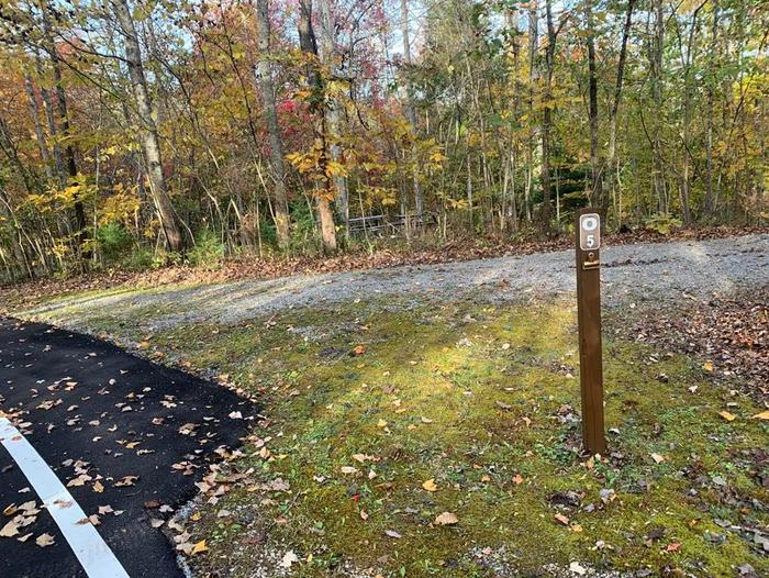 Gravel area with a small brown post near the blacktop road.O-5 has a fire ring, grill, and picnic tables.