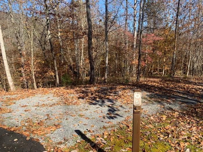 Gravel area with a small brown post near the blacktop road.O-6 has a fire ring, grill and picnic tables.