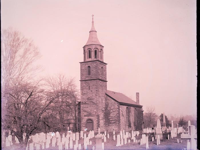 Stone & brick church, with tall steeple and cross, surrounded by gravestones.  St. Paul's Church, which dates to the 1760s, and adjoining historic cemetery, pictured here in the 1930s. 