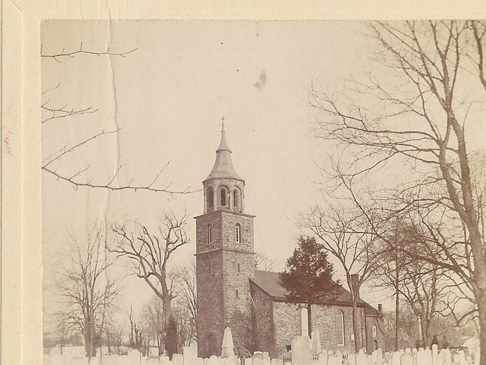 Stone church with steeple, surrounded by gravestones, snow on ground. St. Paul's Church and cemetery, winter scene, mid 20th century. 