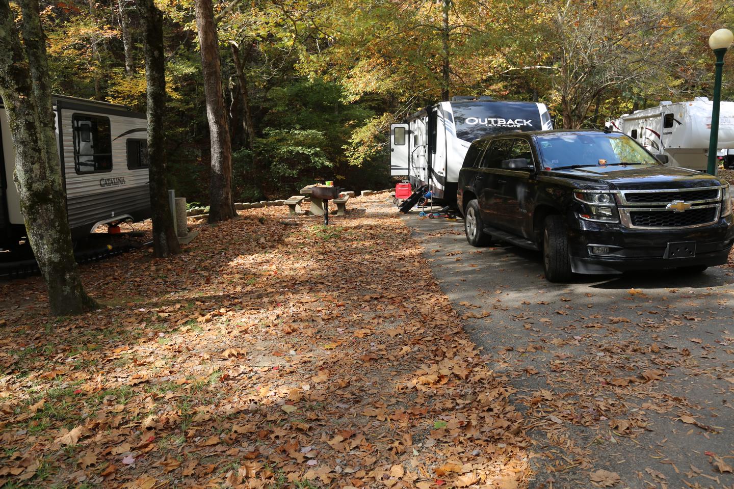 Camper with SUV in front parked on a paved pad with a grill and picnic table to the left, and surrounded by treesCampsite 37