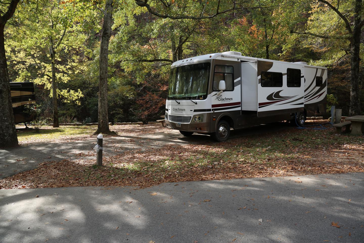 Large RV with side popped out on paved site surrounded by trees, grass and fallen leavesCampsite 41