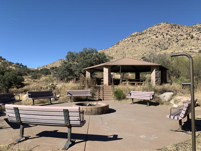 Uncovered fire pit surrounded by benches, with the covered pavilion with picnic table in the background.Reserve the group campground for Big spacious meeting spaces surrounded by designated tent pads.