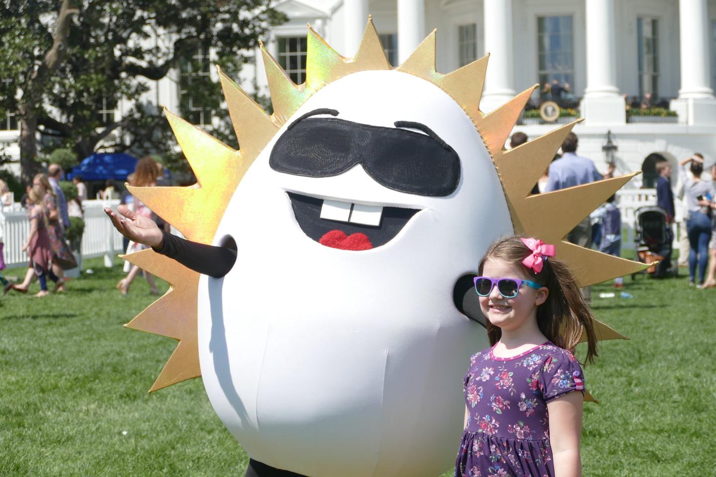 A child posing with a sunshine egg mascotGuests attend the White House Easter Egg Roll, on the South Lawn of the White House. 