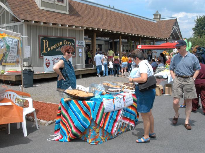 Two people check out a table staffed by a volunteer as part of community event. In the background, a crowd of approximately 20 people are standing by a train station.Each year, the city of Carbondale hosts their annual Pioneer Nights Community Festival, and end-of-summer tradition.