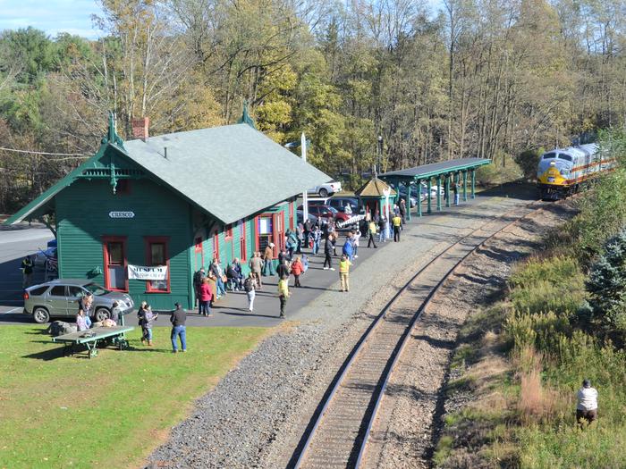 Aerial view of train station with a crowd of approximately 40 people outside waiting for the approaching train.On days of excursions to Cresco Train Station visitors can enjoy a display of antique cars, live music, and artwork.
