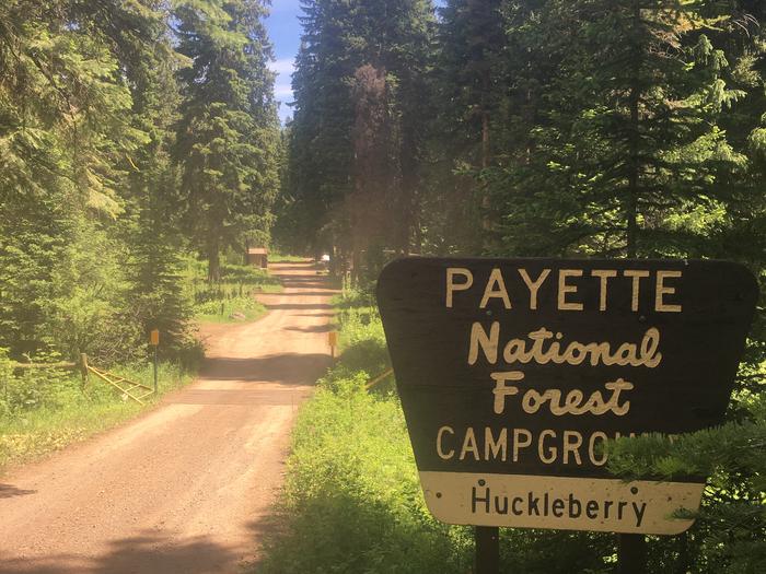 Dirt road with forest alongside and large sign for Huckleberry Campground.Entrance to Huckleberry Campground.
