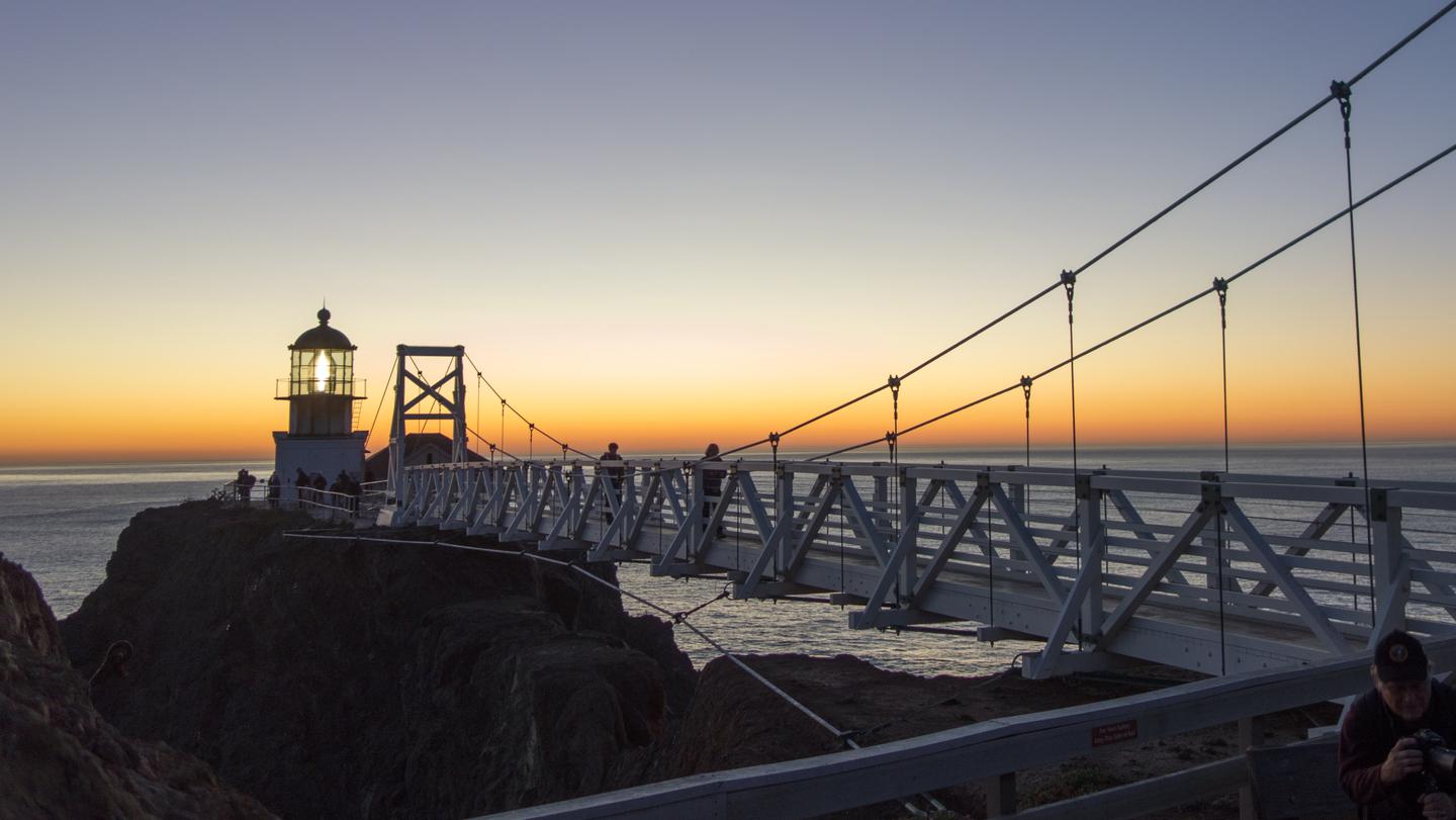White bridge over rocky cliff connects to the Point Bonita Lighthouse. It is dusk, and the glow of the light through the glass lens is visible Point Bonita Lighthouse at sunset