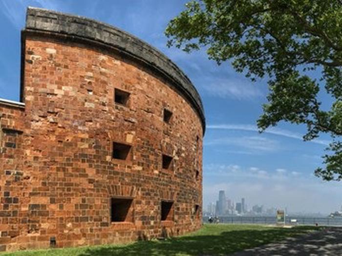 Three story red sandstone eastern wall of Castle Williams with NY Harbor and Skyline in backgroundThe three story, water battery, Castle Williams, designed by its name sake Col J. Williams, to defend New York Harbor