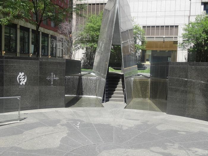 The Ancestral Libation Chamber, with symbols from the African Diaspora and water flowing into two pools. Trees with green leaves and green grass can be seen in the background.The African Burial Ground National Monument Outdoor Memorial in the Spring and Summer.