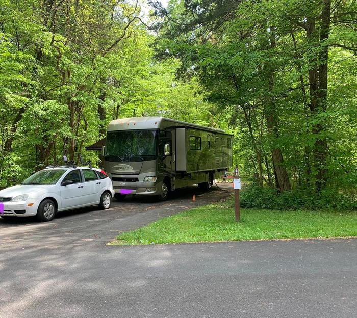 A white car with a brown RV surrounded by green trees.