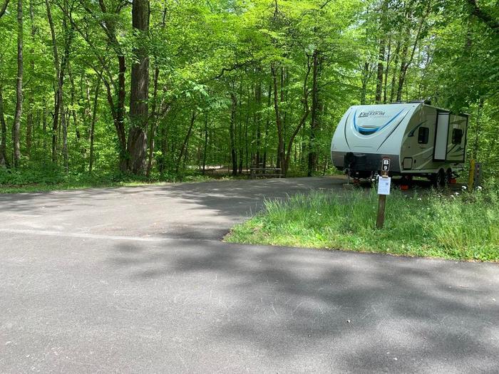 A blacktop surface with a white camper with green trees surrounding it.