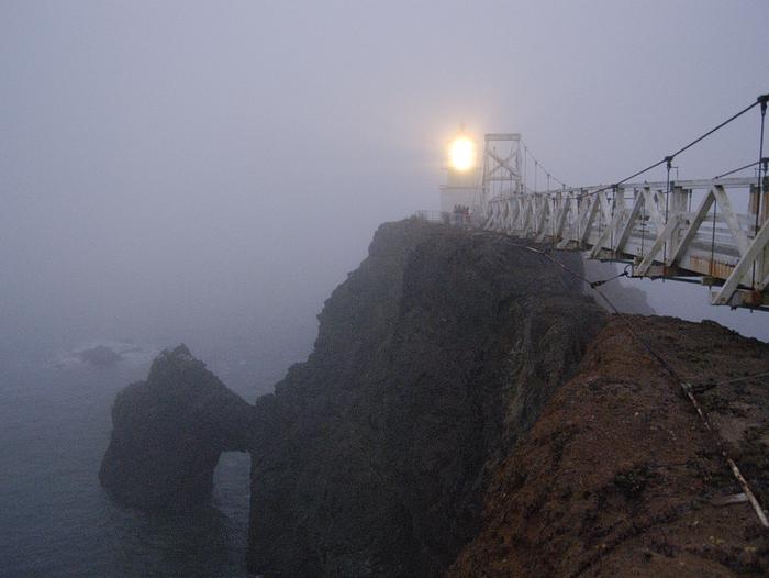 Point Bonita light glows through the fog. Below the lighthouse are dark cliffs and the pillow basalt archway.Point Bonita Lighthouse on a foggy sunset.