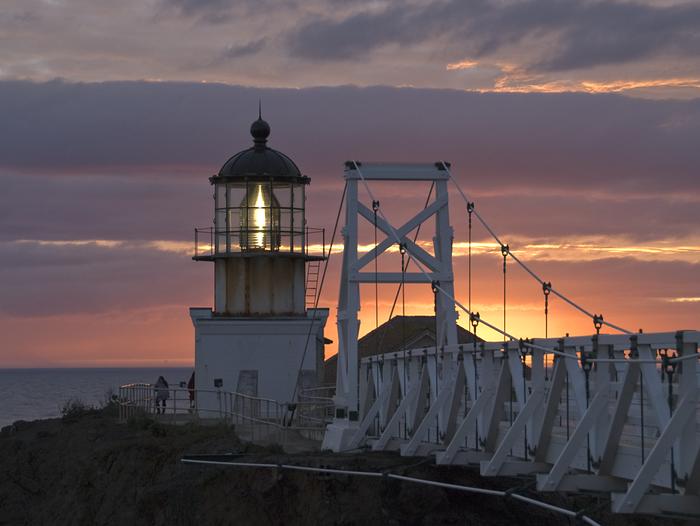 The Point Bonita Lighthouse light shines bright against a darkening sunset sky over the ocean/Point Bonita Lighthouse at sunset on a cloudy evening.