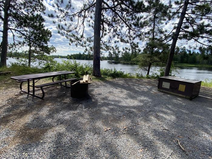 R26 - Sunrise Point, view looking out from campsite with a picnic table, bear box, and a fire ring with wood on grate.R26 - Sunrise Point campsite on Rainy Lake