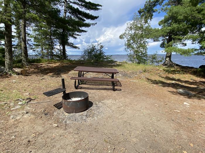 R62 - Krantz Point, view looking out from campsite of the fire ring and picnic table with water in the background.R62 - Krantz Point campsite on Rainy Lake