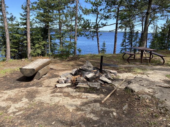 R63 - Loon Bay, view looking out from campsite with a fire ring, picnic table, and a wood bench with water in the background.R63 - Loon Bay campsite on Rainy Lake