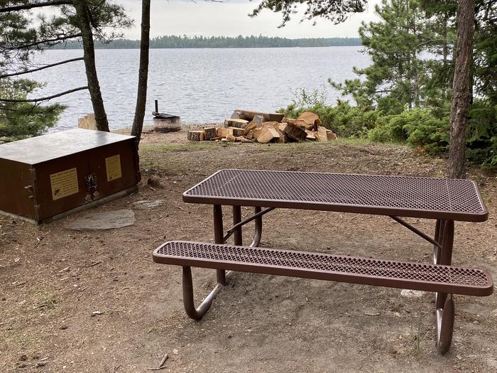 R64 - Mio Beach, view looking out from campsite with a picnic table, bear box, wood pile, and fire ring with water in the background.R64 - Mio Beach campsite on Rainy Lake