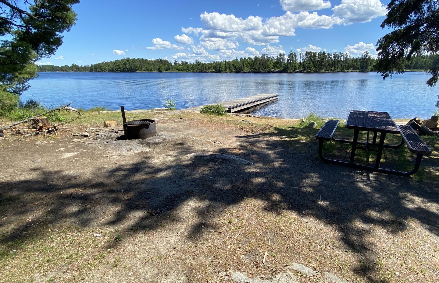 R74 - Rainy Lake Group, view looking out from campsite of a fire ring and picnic table with one of the boat docks in the background of water.View looking out from campsite.