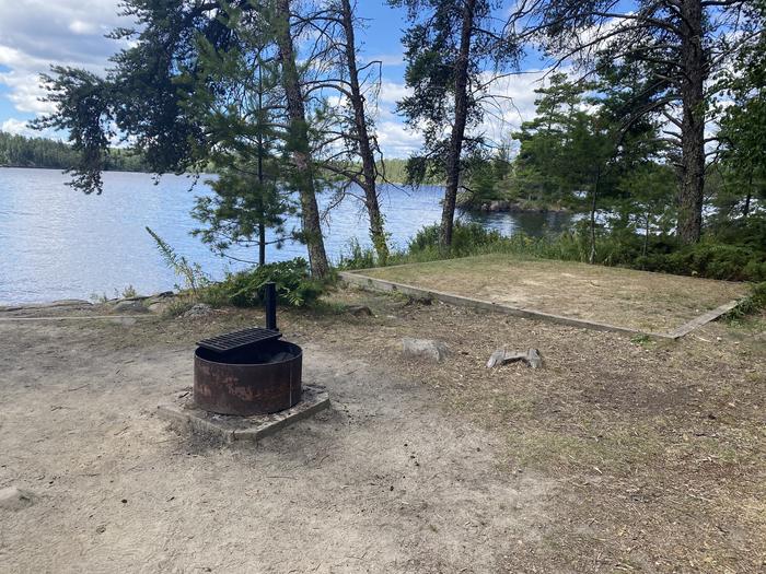 R104 - Saginaw Bay West, view looking out of campsite with a fire ring and tent pad with water in the background.R104 - Saginaw Bay West campsite on Rainy Lake
