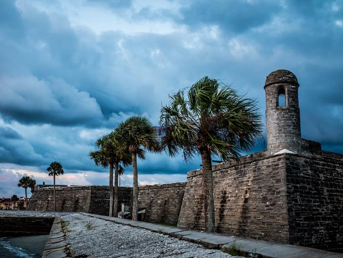 A gray stone structure with high walls and a tall bell tower dominate the image. Palm trees are in the foreground with a gray sky overhead. Castillo de San Marcos