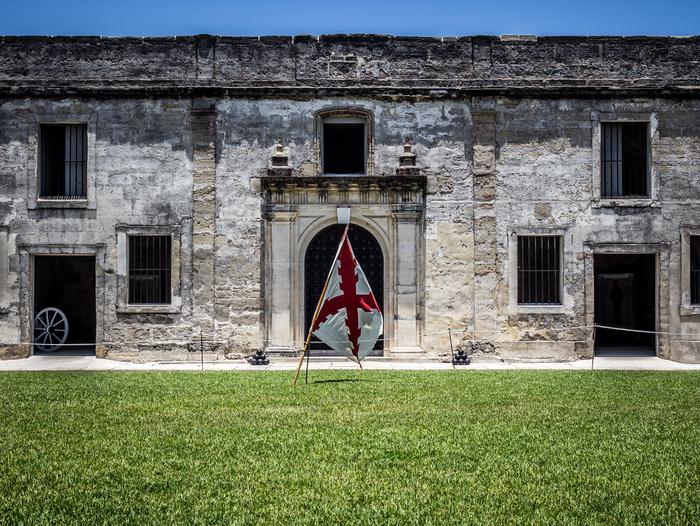 Gray stone wall with several door and window openings. There is green grass in the foreground with a white flag with a red diagonal cross hanging on a wooden pole. Castillo de San Marcos Interior Courtyard