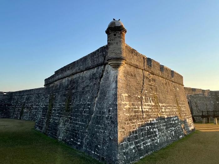 Gray fort wall with diamond-shaped bastion dominating the photo. Blue sky above the fort and green grass below. Castillo de San Marcos