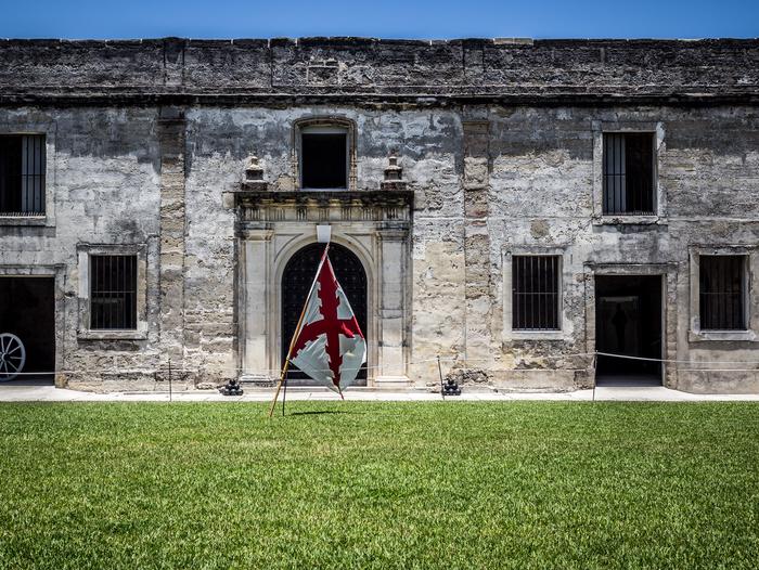 Gray stone wall with door and window openings. Green grass in the foreground with a white flag with a red diagonal cross hanging on a wooden pole. Interior Courtyard of Castillo de San Marcos