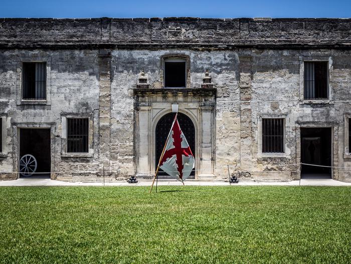 Gray stone wall with several door and window openings. Blue sky at top of photo, green grass in foreground. White flag with red diagonal cross on wooden pole in middle of photo. Interior Courtyard of Castillo de San Marcos.