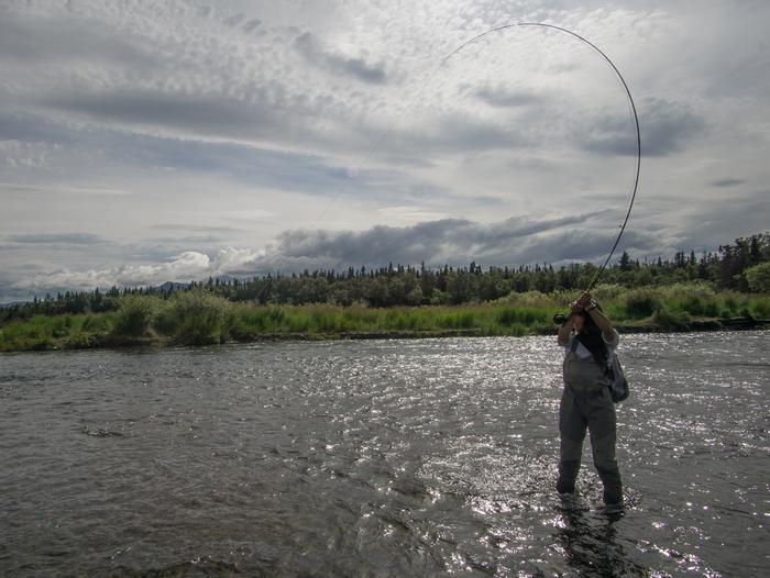 An angler stands in a shallow river holding up a fishing rod bent with the weight of a fish on the lineThe Brooks River offers opportunities for fishing, photography, and wildlife viewing.