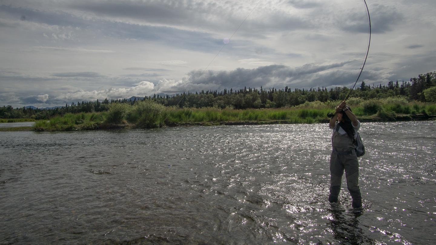 An angler stands in a shallow river holding up a fishing rod bent with the weight of a fish on the lineThe Brooks River offers opportunities for fishing, photography, and wildlife viewing.