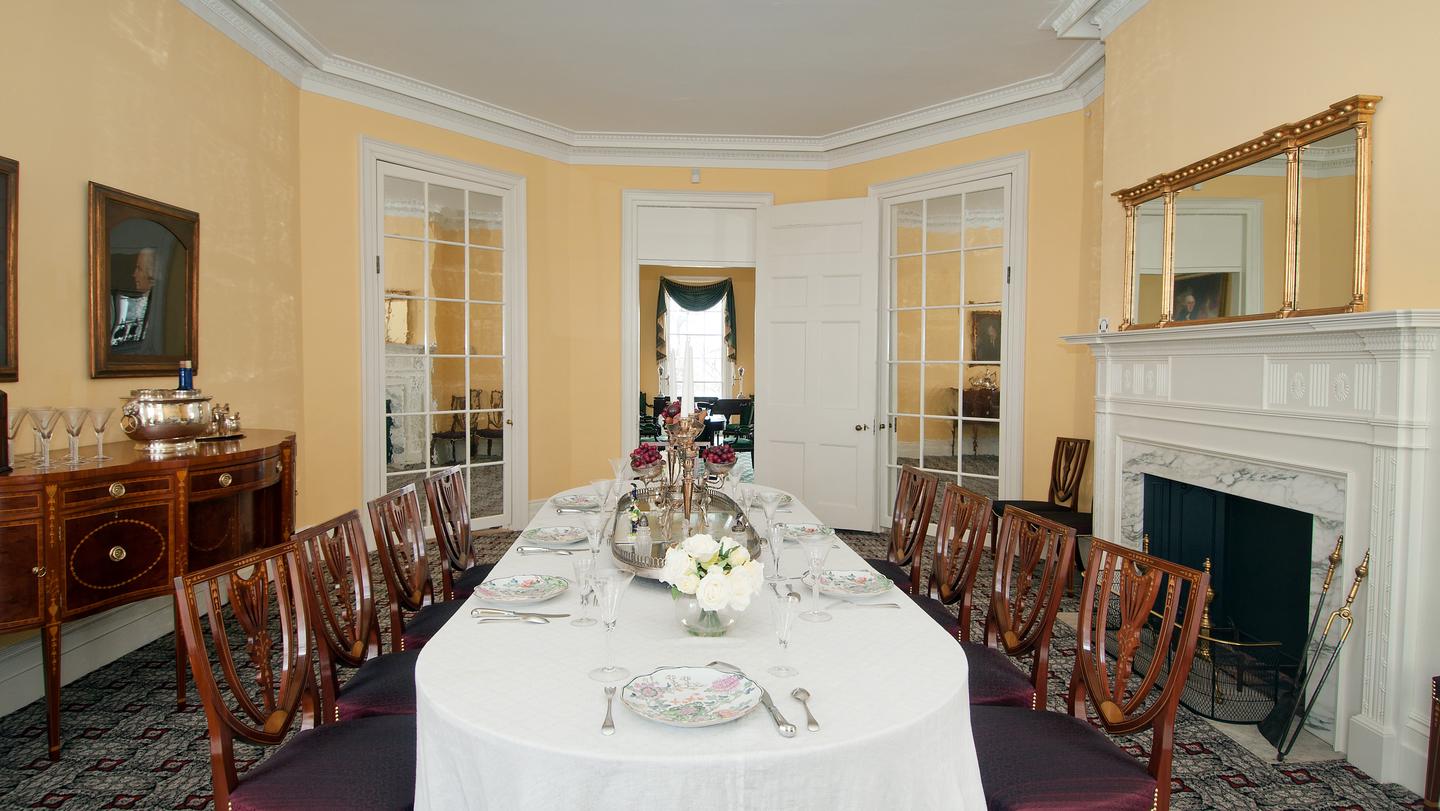 A dining table lined with chairs in a yellow room.The Hamilton Grange dining room is the formal location for Hamilton's dinner parties in the country.