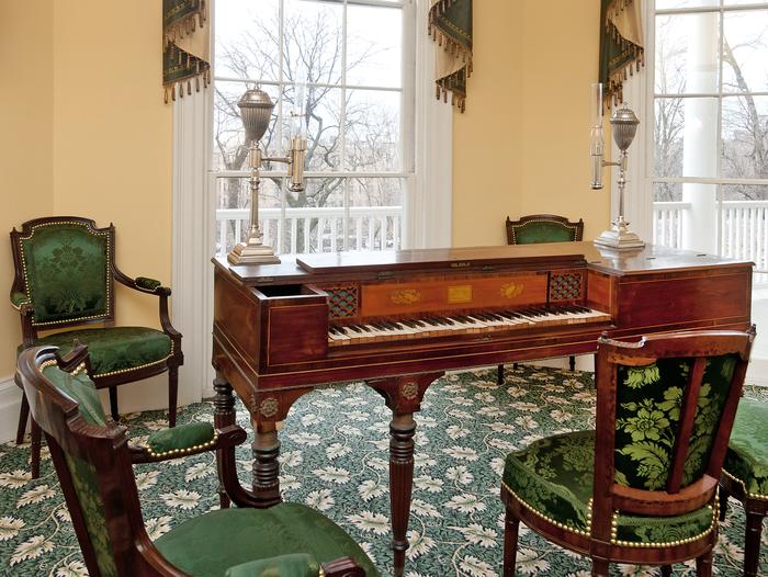 A wooden piano surrounded by green upholstered chairs. The piano of Angelica Hamilton is on display in the Hamilton Grange parlor.