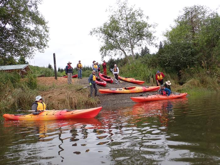 A group of people wearing life jackets stand on a river bank among brightly colored kayaks. A ranger stabilizes a floating kayak as a paddler positions themselves inside. Nearby, another paddler in a warm-colored kayak pauses in the water.Rangers assisting paddlers into the river at the beginning of a paddle tour