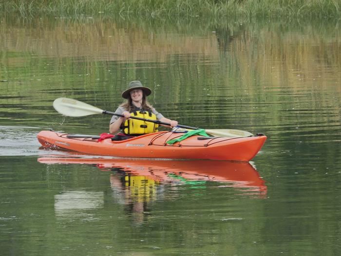 A ranger in a yellow life jacket paddles an orange kayak on rippling green water.Ranger Esther leading a kayak program