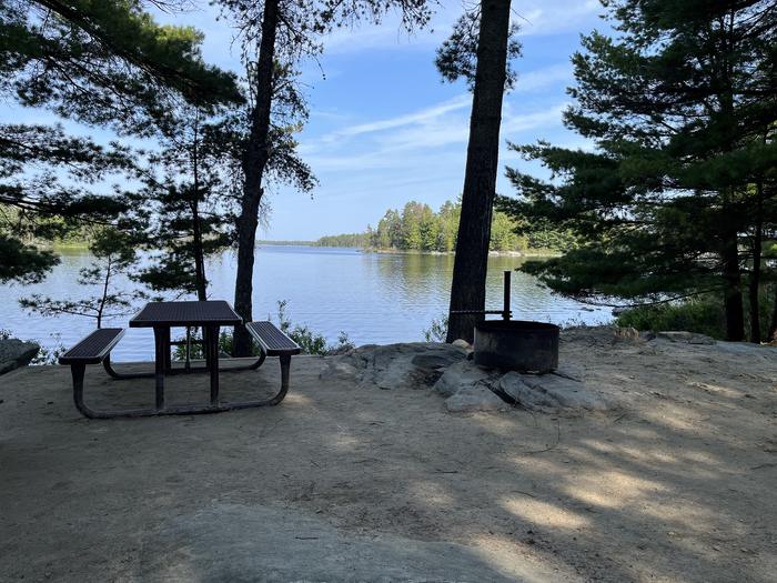 R59 - Finlander Island, view of the fire ring and picnic table overlooking the water through a couple of trees.R59 - Finlander Island campsite on Rainy Lake