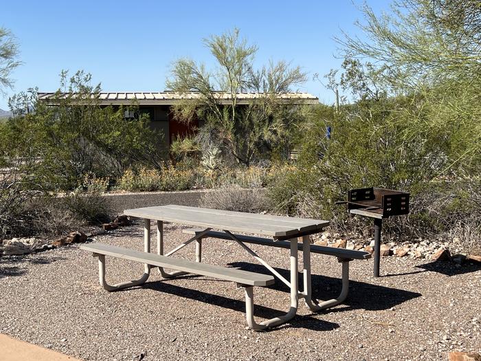 A picnic table sits near a grill and desert vegetation with the bathroom behind.Each site has a picnic table and grill.